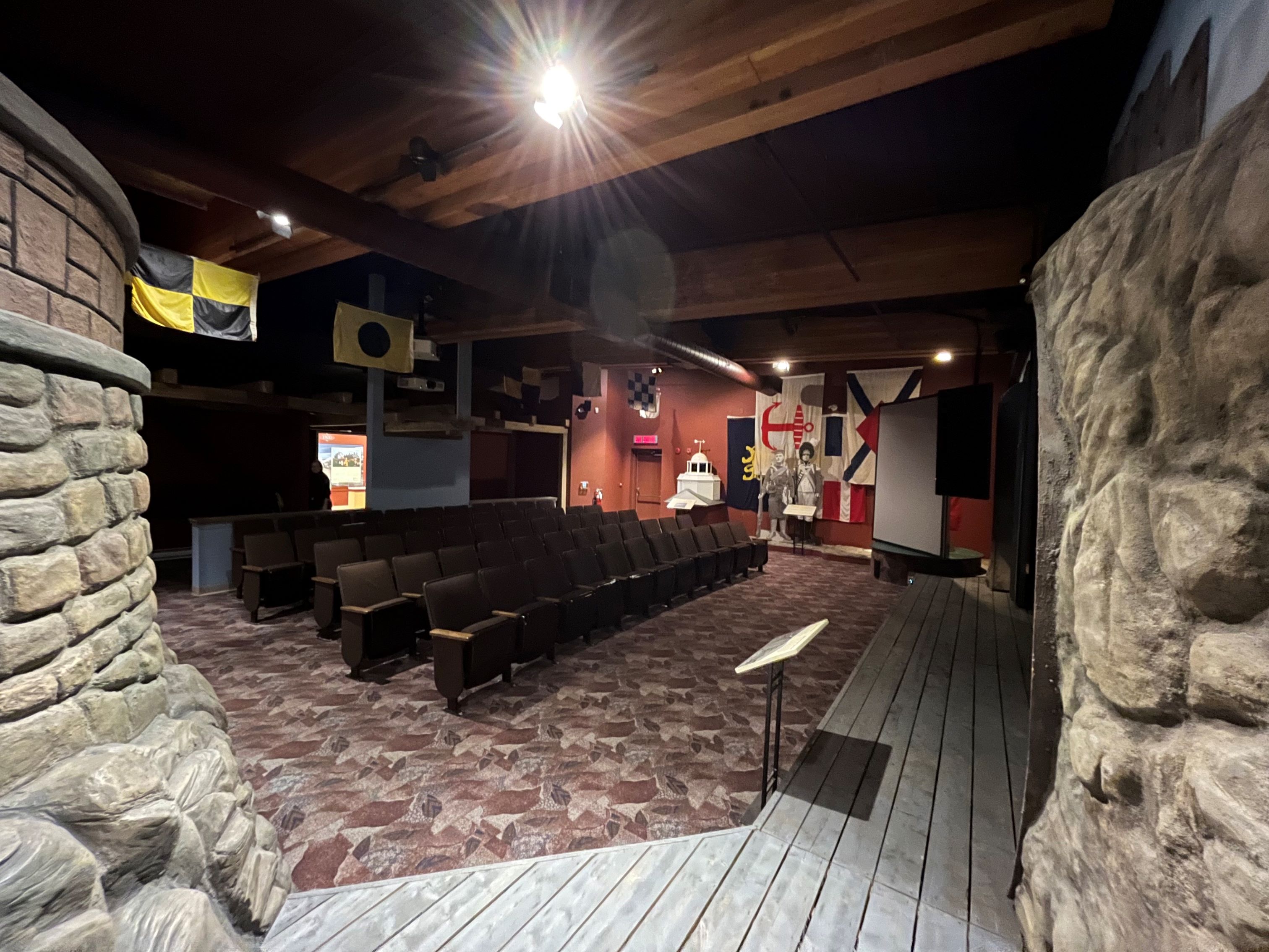 Four rows of theatre seating are seen from the side stage perspective. Several flags are hanging from the ceiling.