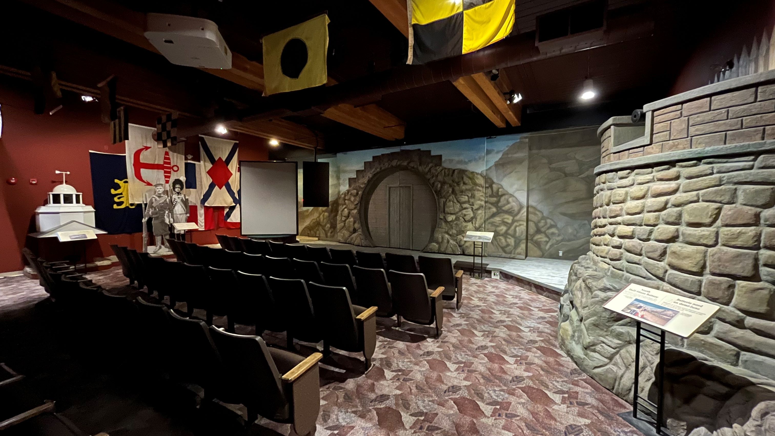 Four rows of theatre seating are seen from behind with the stage in full view in front of them. The stage has a painted backdrop that illustrates a stone wall with a wooden door and several flags are hanging from the ceiling.
