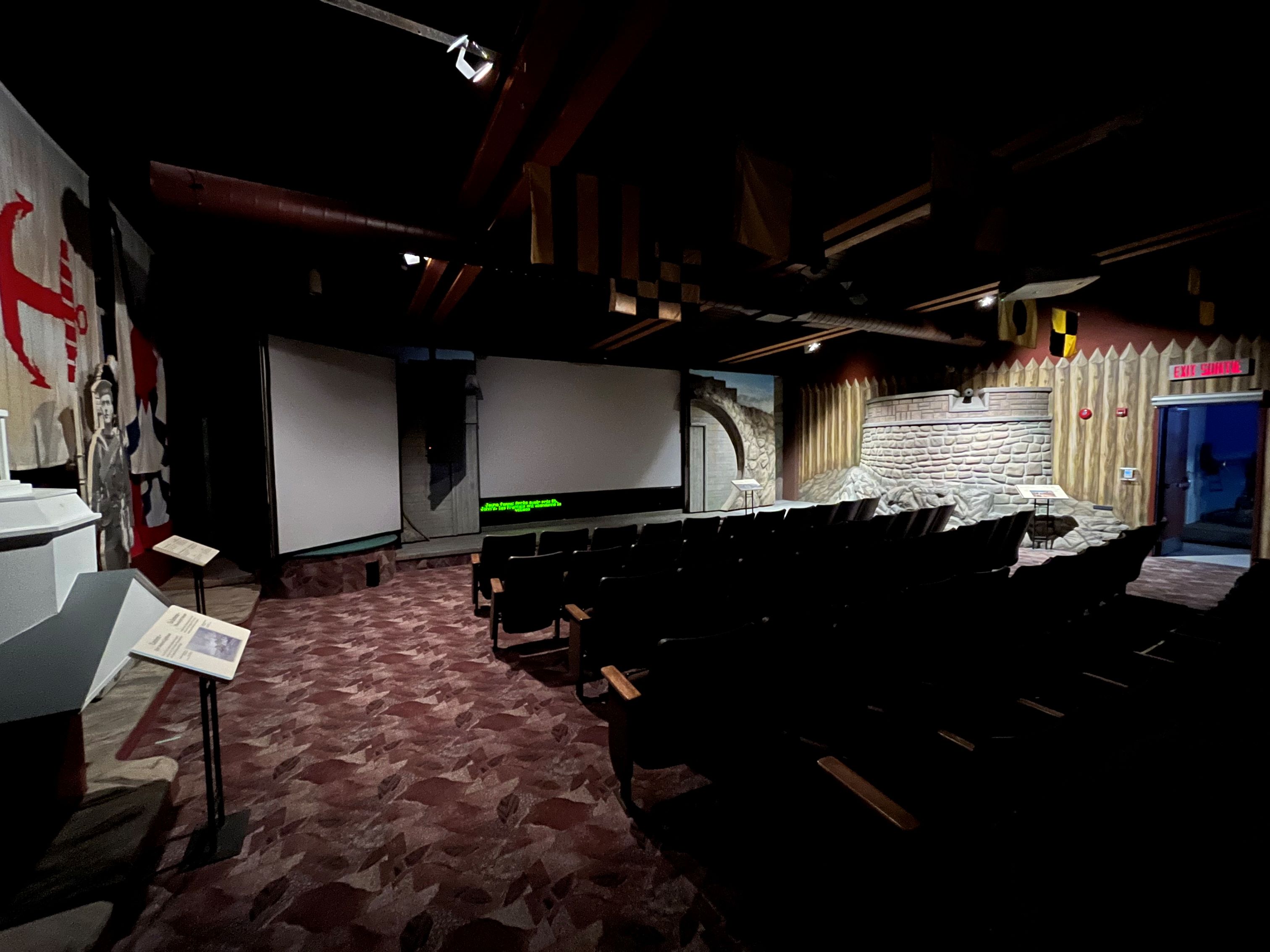 Five rows of theatre seating are seen from behind with the stage in full view in front of them. Two projectors are lowered at the stage and the lights in the room are dimmed.