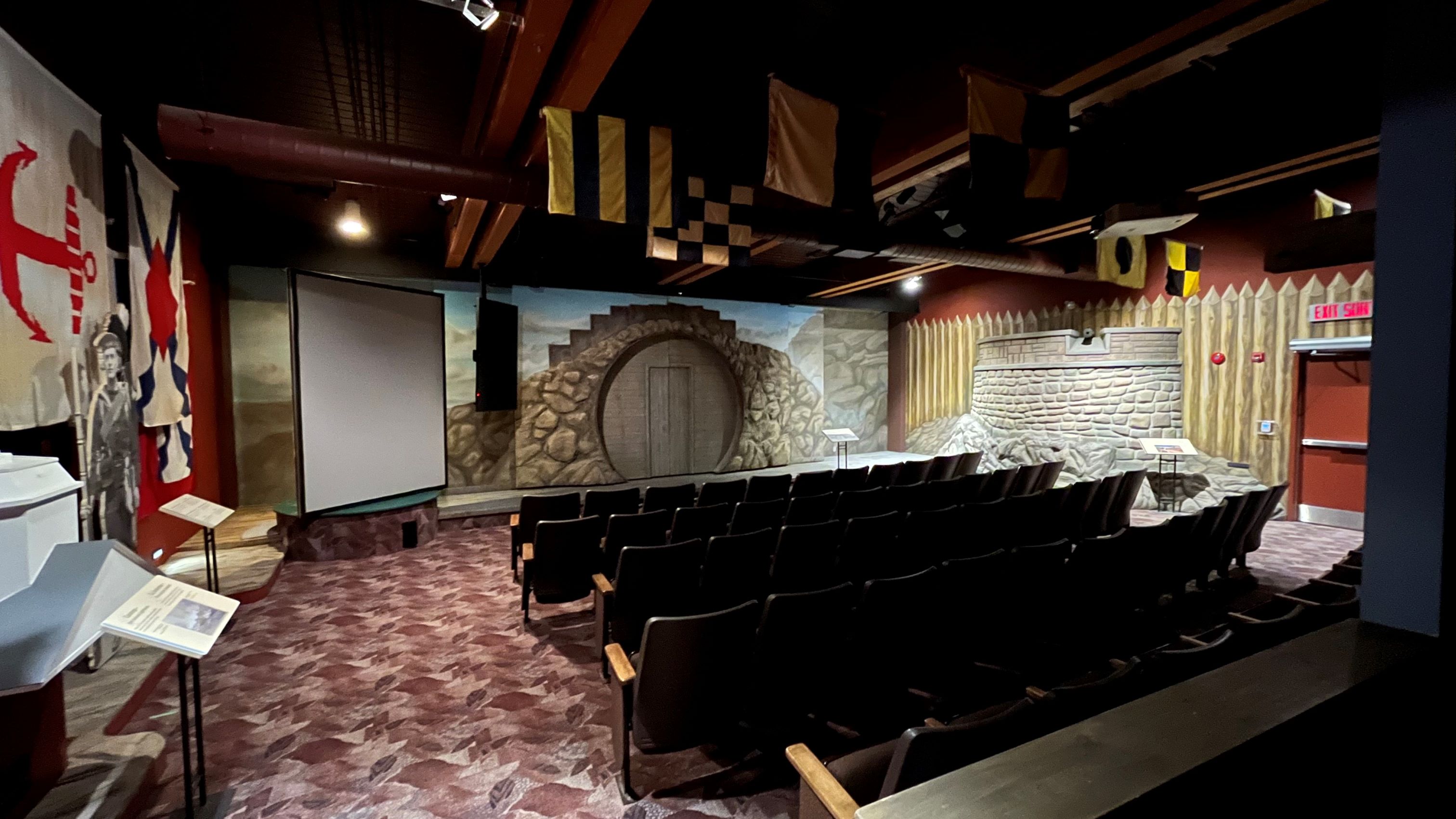 Five rows of theatre seating are seen from behind with the stage in full view in front of them. The stage has a painted backdrop that illustrates a stone wall with a wooden door and several flags are hanging from the ceiling.