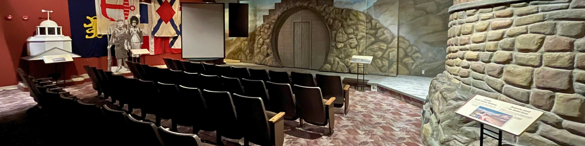 Four rows of theatre seating are seen from behind with the stage in full view in front of them. The stage has a painted backdrop that illustrates a stone wall with a wooden door and several actual flags are hanging from the ceiling at the left.