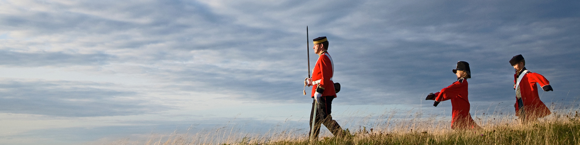 a Parks Canada employee in historic uniform, leads two children wearing large historic uniforms