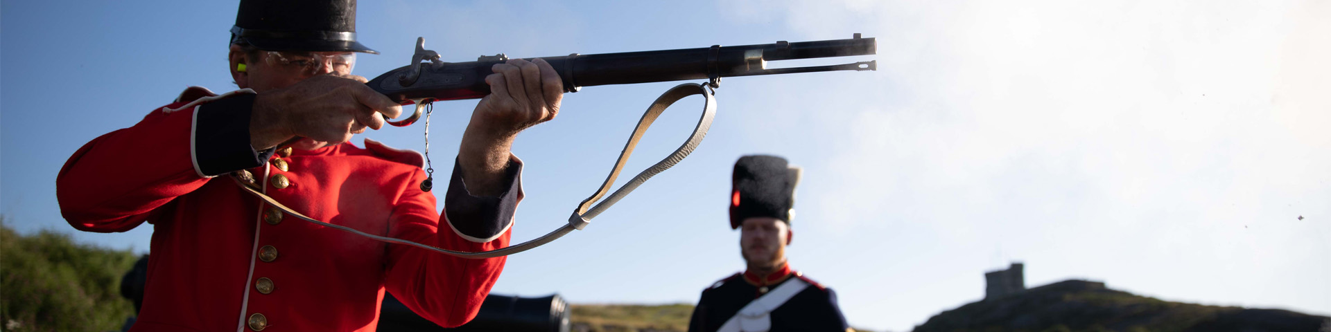 two individuals in historic military uniform; one is firing a musket and the other stands on guard