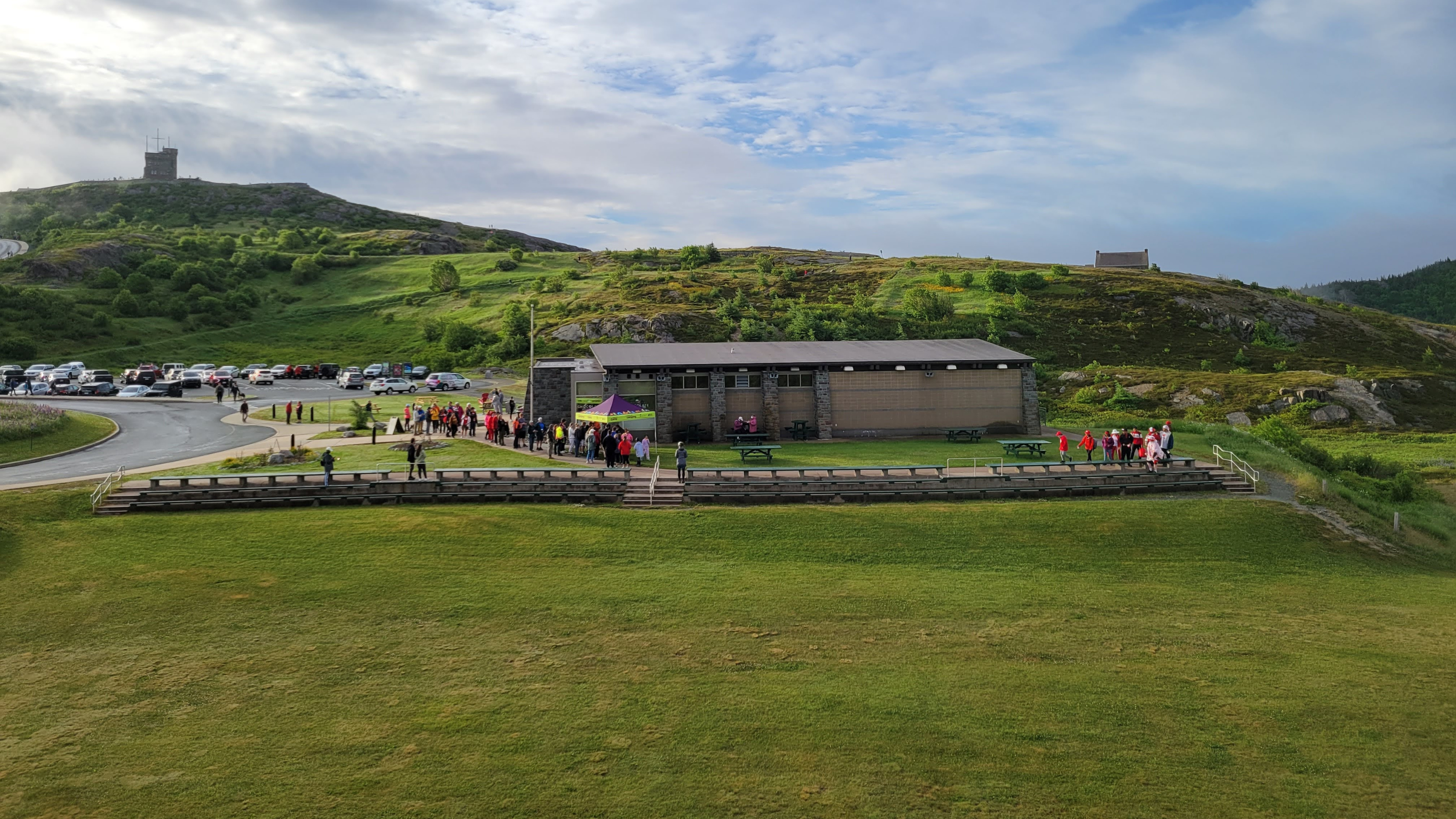 A wide photo shows a big open field with a stone building in the centre behind it and three levels of wooden bench seating along the field’s edge.