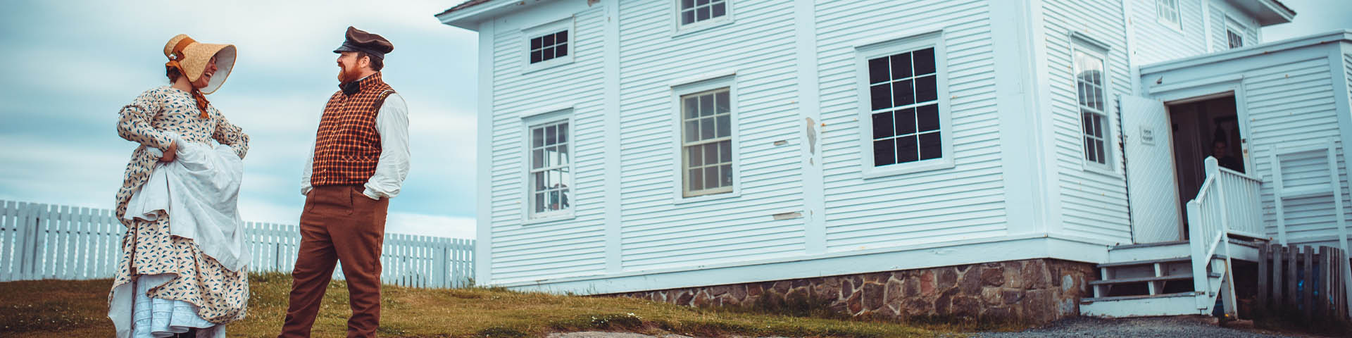 two individuals in historic costume, laughing outside of a white, wooden house.