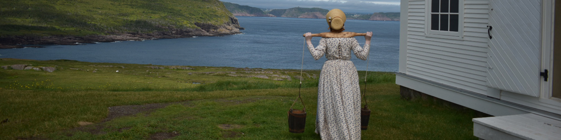 a woman in historic costume carrying water looks at a  a coastal view by a lighthouse