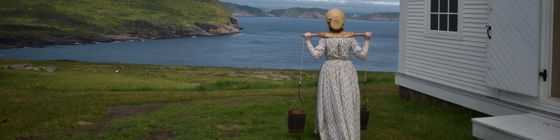 a woman in historic costume carrying water looks at a a coastal view by a lighthouse