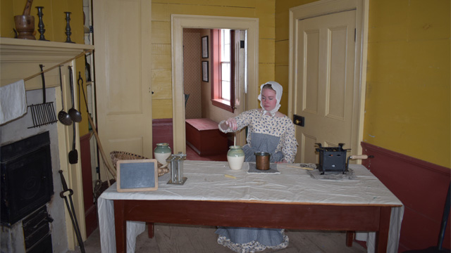a woman in historic costume sitting at a wooden table, making candles 