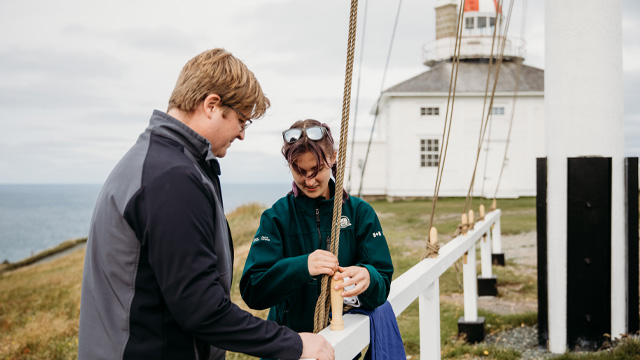 Un employé de Parcs Canada aide une personne à préparer un drapeau sur des cordes, avec un phare en arrière-plan.