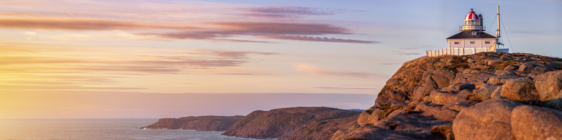 a white, square, lighthouse on a rocky cliff overlooking the ocean under a colourful sunrise