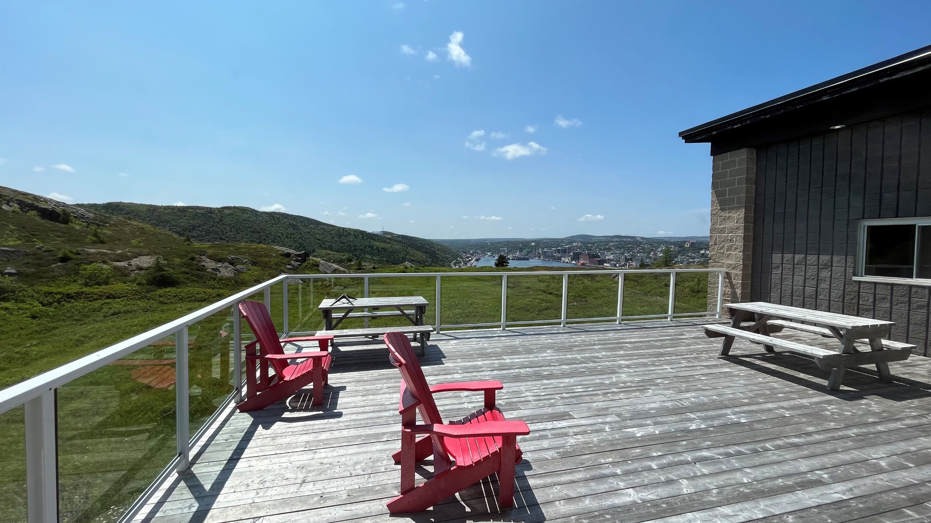 A wooden deck is seen with two red chairs and a wooden picnic table with a view of hills and a cityscape in the background.