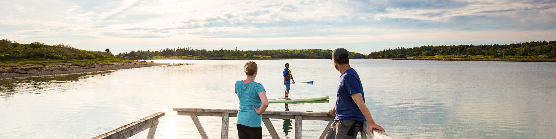 Deux personnes sont debout sur un quai et regardent quelqu'un faire du paddle board pendant que le soleil se couche.