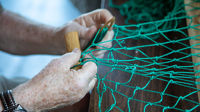 Deux mains tenant des outils en bois pour fabriquer un filet de pêche vert.