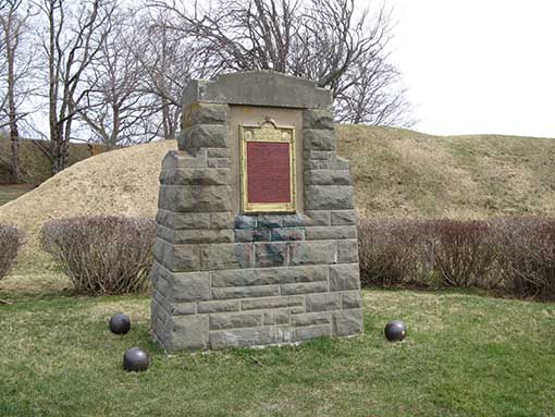 A stone monument with a maroon plaque. Four cannon balls rest on the grass at four corners of the base.