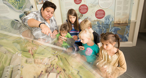 Une femme et un groupe d’enfants regardent une maquette du fort sous un plexiglas.”><figcaption class=