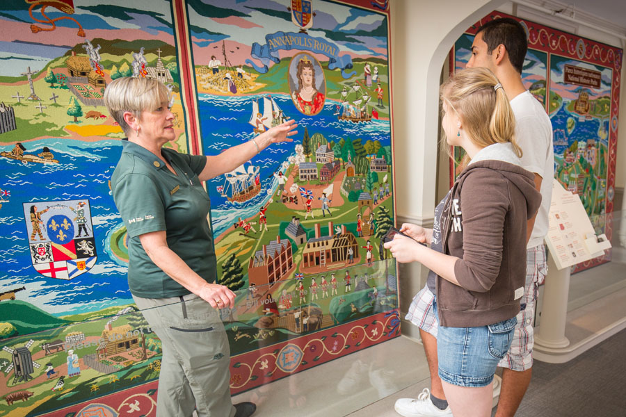 Two visitors listen to a Parks Canada tour guide explaining the large and colourful heritage tapestry covering the wall.