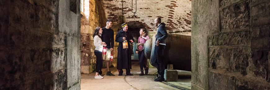 Un guide-interprète costumé tient une lanterne et s’adresse à un groupe de visiteurs à l’intérieur d’une petite pièce en briques dotée d’un canon.