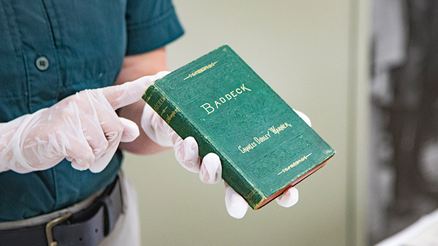 Hands wearing white gloves hold a book entitled Baddeck