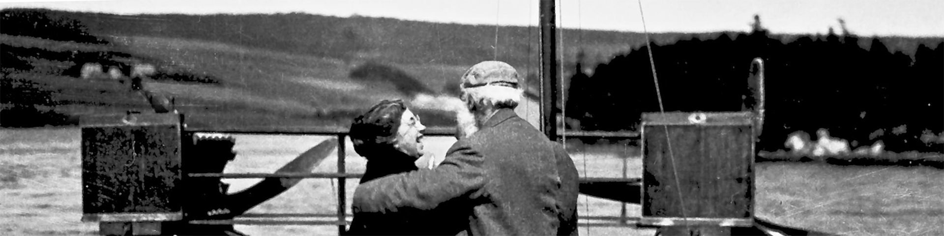 An historical photo of Mabel smiling up at Alexander Graham Bell while standing on a dock, they both have their backs towards the camera. 