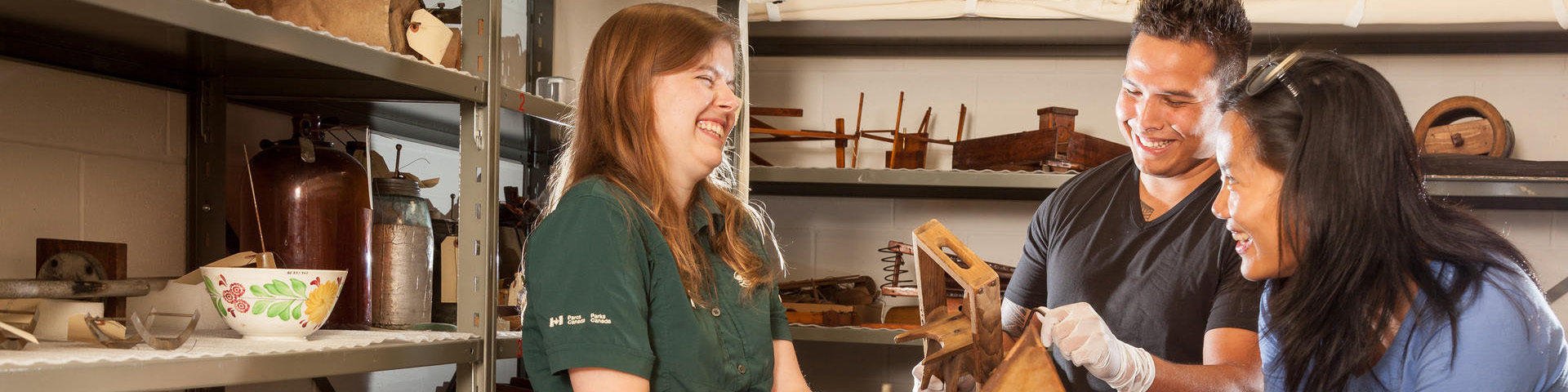 A uniformed Parks Canada interpreter and two visitors smile at each other standing in an exclusive collections room while learning about artifacts and inventions