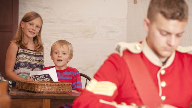 Two young visitors smile and hold a “Citadel Adventures” booklet while looking at an interpreter in soldier’s uniform at Halifax Citadel National Historic Site.