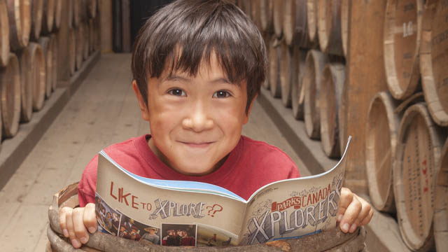 A young boy stands inside a barrel in the powder magazine, smiling and looking up from an Xplorers booklet at Halifax Citadel National Historic Site.