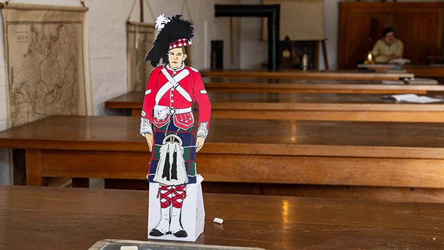 Un soldat en papier du 78e régiment des Highlanders est posé sur une table en bois dans la salle de classe. On peut voir un interprète historique représentant le maître d’école assis à son bureau à l’arrière-plan.