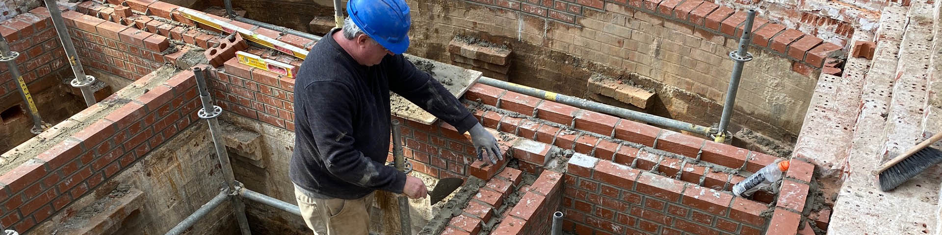 Standing in a small, uncovered brick chamber, a worker lays new bricks for the chamber walls.