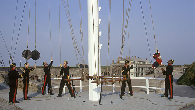 Six soldiers in blue pull the ropes to raise flags on the signal mast.