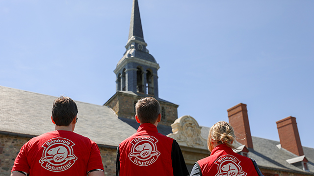Trois personnes regardent une tour d'horloge. Elles portent des vestes de bénévole rouges avec le logo de Parcs Canada.