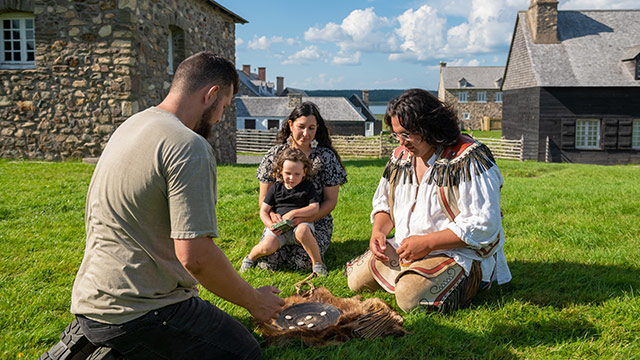 A family with a small child sit in the grass with traditionally dressed Mi’kmaw interpreter