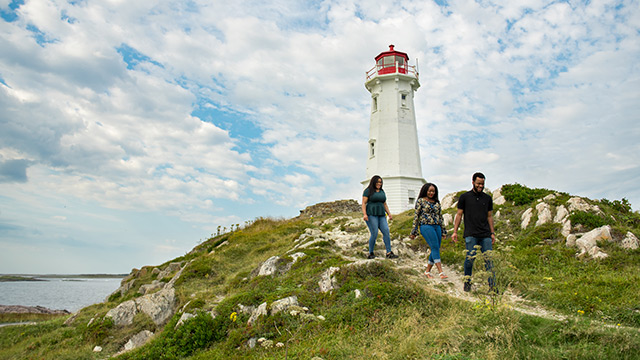 Trois personnes marchent sur un sentier de randonnée avec un phare historique en arrière-plan par une journée ensoleillée.