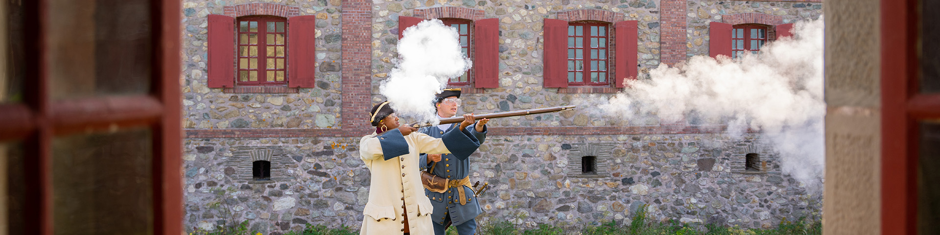 a visitor fires a musket with a costumed solider, there is smoke in the air to the right of them.