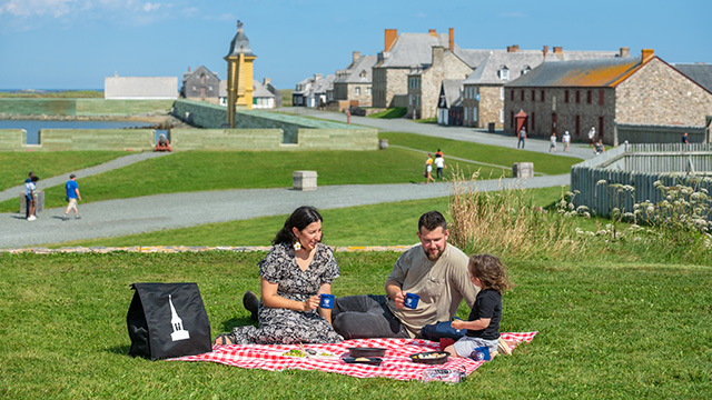A family of three sits on the grass while they have a picnic at the Fortress of Louisbourg 