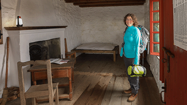 A person stands next to a door inside a room of an 18th-century building while they hold a sleeping bag and have a backpack on their back,