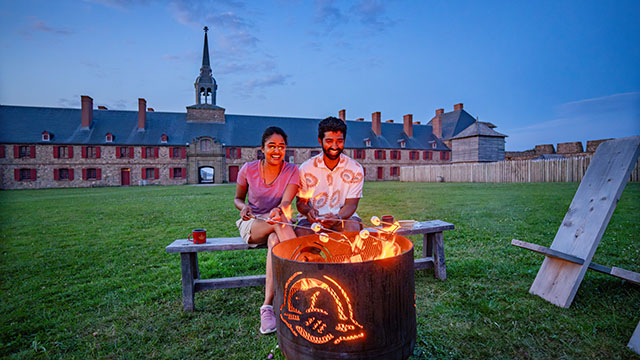 Deux personnes font rôtir des marshmellos sur un foyer au lieu historique national de la Forteresse-de-Louisbourg. 
