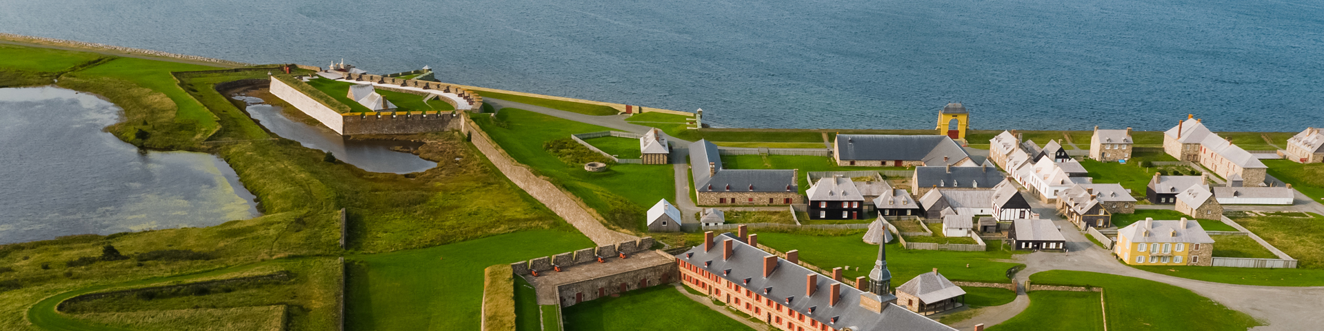 Une vue aérienne du lieu historique national de la Forteresse de Louisbourg, montrant plusieurs bâtiments avec l'océan en arrière-plan.