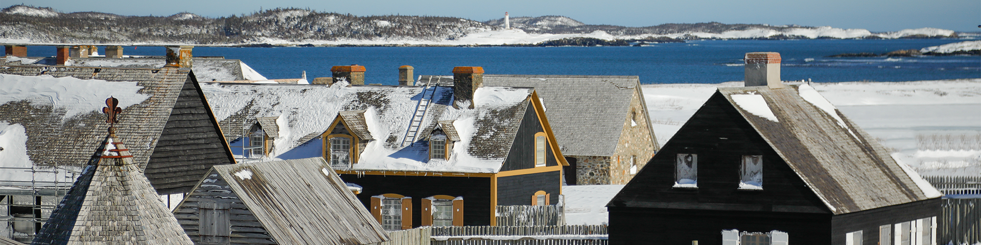 Vue sur les toits enneigés du Lieu historique national de la Forteresse-de-Louisbourg. Au loin, on aperçoit un phare. 