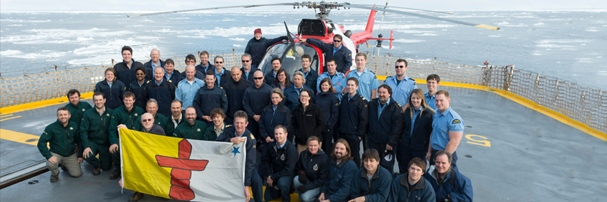 A group of people holding a Nunavut flag. A helicopter is in the background.