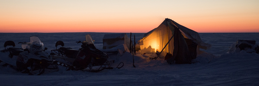 A tent and two snowmobiles at sunset.