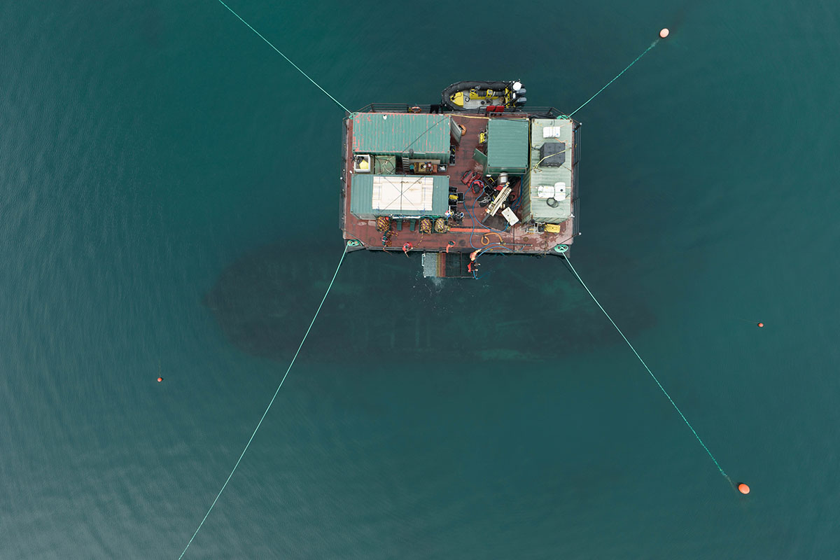 Aerial view of a floating barge anchored in water, with green-roofed shipping containers and equipment, secured by lines to surrounding buoys.