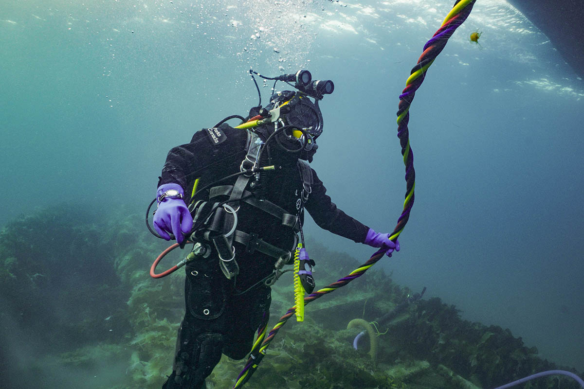 Scuba diver underwater holding an umbilical line, wearing full diving gear with marine vegetation and submerged shipwreck in the background.