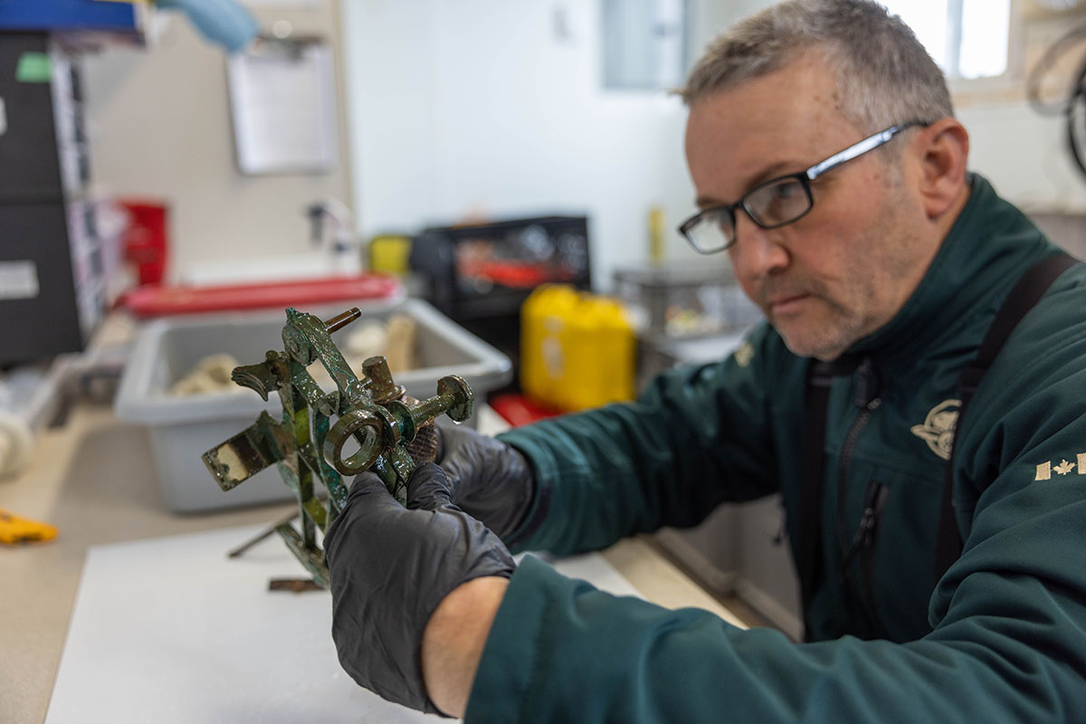 Person wearing gloves and a Parks Canada uniform in a lab with equipment and containers in the background examining a sextant artifact recovered from a shipwreck.