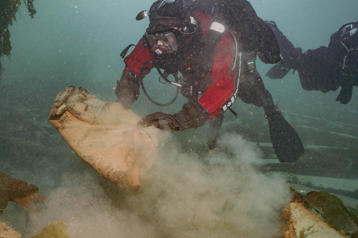 Scuba diver in red and black gear lifting a sediment-covered folded coat from the seafloor, with another scuba diver and marine vegetation in the background.