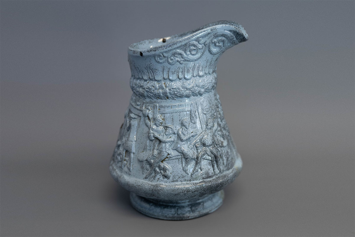 Ceramic pitcher with a spout, decorated with raised relief patterns and scenes depicting figures engaged in various activities.