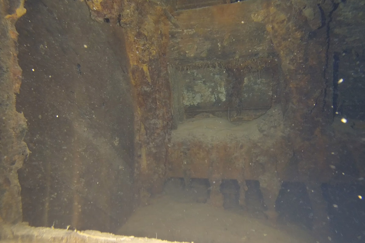 Underwater view of a rusted and decayed shipwreck interior, with corroded walls and ceiling covered in marine growth.