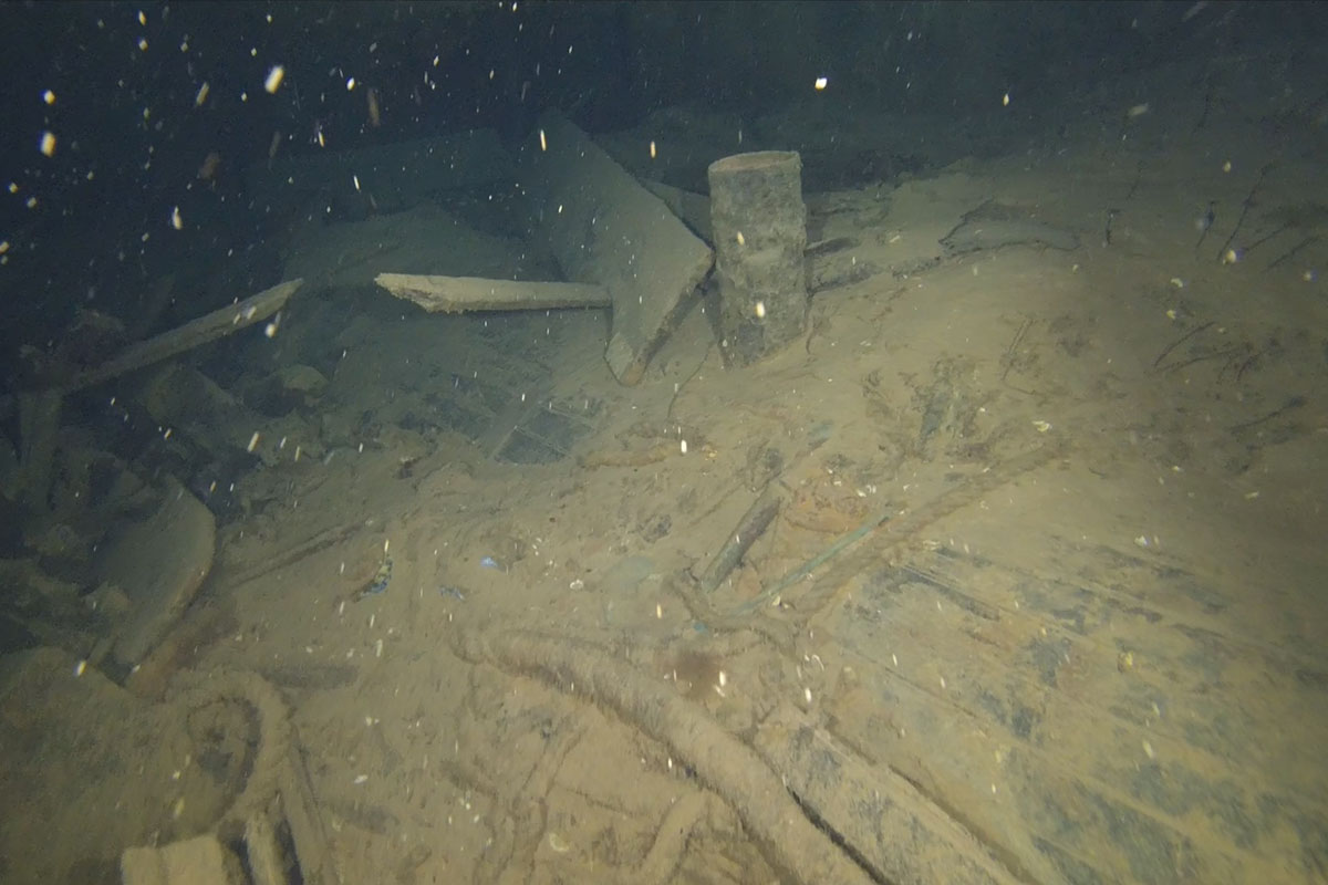 Underwater scene with scattered debris inside a wreck, including wood and metal pieces covered in sediment, surrounded by floating particles.