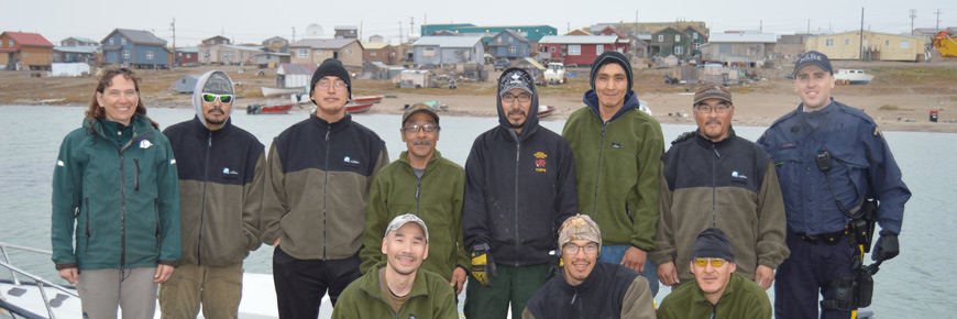 Eleven people standing on the shore, Gjoa Haven is in the background.