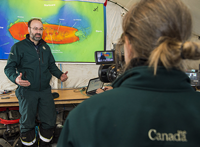 A Parks Canada employee filming another Parks Canada employee standing in front of an image of the Erebus shipwreck and talking to the camera.  