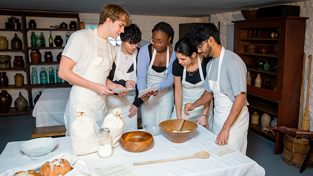 A group in aprons stands over a table with bowls and ingredients. 